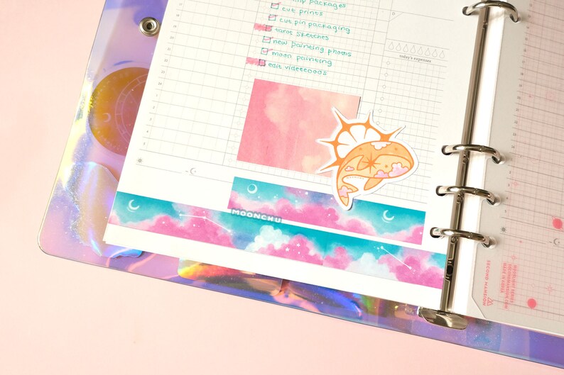 Op de afbeelding: Een witte planner met een transparante plastic hoes met een holografisch ontwerp. De planner heeft een ringband en een pagina met een roze aquarel achtergrond. Een sticker van een walvis met een ster op zijn rug is op de pagina. De pagina heeft ook een decoratieve washi tape met een roze en blauwe hemel en sterren ontwerp.