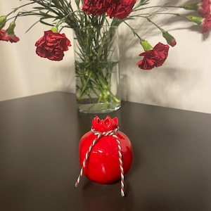 May include: A glossy red ceramic pomegranate ornament with a decorative red flower top and a twisted green and white rope. A clear glass vase with red carnations is in the background.