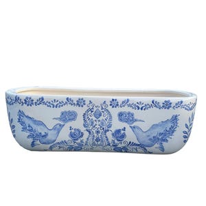 Può includere: Una fioriera rettangolare in ceramica bianca con un design floreale e di uccelli blu e bianco. La fioriera presenta un motivo dettagliato di fiori, nuvole e uccelli, creando un aspetto decorativo e artistico. Ideale per uso interno o esterno.