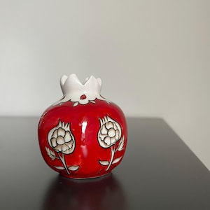 May include: A red ceramic pomegranate-shaped vase with a white crown and floral designs. The vase has a glossy finish and features two white and beige flower illustrations. The vase is a decorative item.