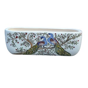 Op de afbeelding: Een witte keramische plantenbak met een rechthoekige vorm. De plantenbak heeft een handgeschilderd ontwerp van twee pauwen die op een tak zitten met groene bladeren en rode bessen. De pauwen hebben blauwe en groene veren.