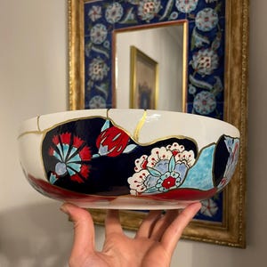 May include: A white ceramic bowl with a navy blue and red floral design, accented with gold kintsugi repairs. The bowl is held up in front of a framed mirror with a blue floral tile border.