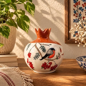 Könnte beinhalten: Eine Keramikvase in Granatapfelform mit glänzend orangefarbenem Oberteil und weißem Unterteil. Der Körper zeigt eine handgemalte Illustration eines Gimpels, der auf einem Ast mit grünen Blättern und roten Beerenclustern sitzt.