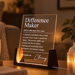 Può includere: Una targa rettangolare in acrilico trasparente inserita in una base di legno scuro con illuminazione LED calda integrata. La targa presenta un testo stampato in bianco che definisce il termine Difference Maker, seguito dal nome Cherry in corsivo.