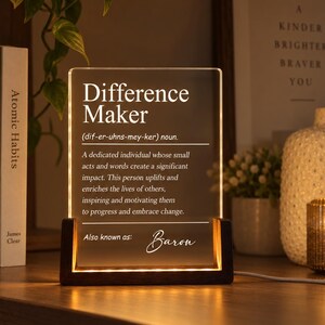 Può includere: Una targa rettangolare in acrilico trasparente con base in legno scuro, illuminata dal basso. Il testo recita: Difference Maker (dif-er-uhns-mey-ker) noun. A dedicated individual whose small acts and words create a significant impact. Also known as: Baron.