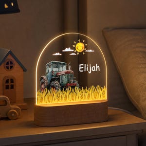 May include: An arched acrylic night light with a wooden base, featuring a printed illustration of a tractor in a field, a smiling sun, and the name Elijah. The lamp emits a warm glow, illuminating the tractor, wheat, and the name in white text.