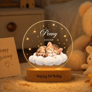 May include: A circular acrylic night light with a wooden base featuring the text Happy birthday. The acrylic panel displays a sleeping tabby kitten on a cloud, surrounded by stars, the name Penny, and the date 21.09.2026 in white script.