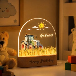 May include: An arched acrylic night light featuring a green tractor in a yellow wheat field, a smiling sun, and the name Gabriel. The light is set in a wooden base with the text Happy Birthday engraved on the front, illuminated with a warm glow.