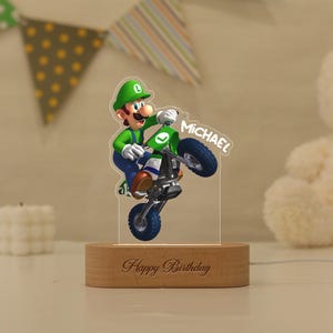 May include: A clear acrylic night light featuring a printed image of Luigi riding a dirt bike, with the name Michael printed in white. The acrylic piece is mounted on a light-coloured wooden base engraved with the text Happy Birthday.