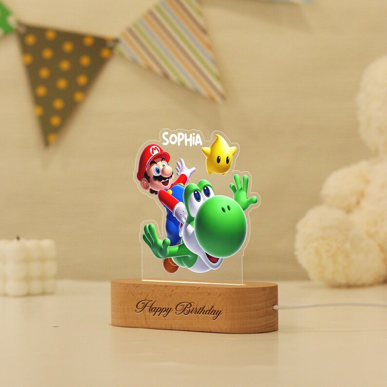 May include: A custom acrylic night light featuring a colourful graphic of Mario riding Yoshi with a yellow star. The name SOPHIA is printed at the top. The light is mounted on an oval wooden base engraved with the text Happy Birthday.