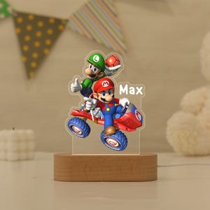 May include: A custom acrylic night light featuring a colourful graphic of Mario driving a red kart with Luigi standing behind him. The name Max is printed in white text on the right side. The acrylic piece is mounted on a smooth oval wooden base.