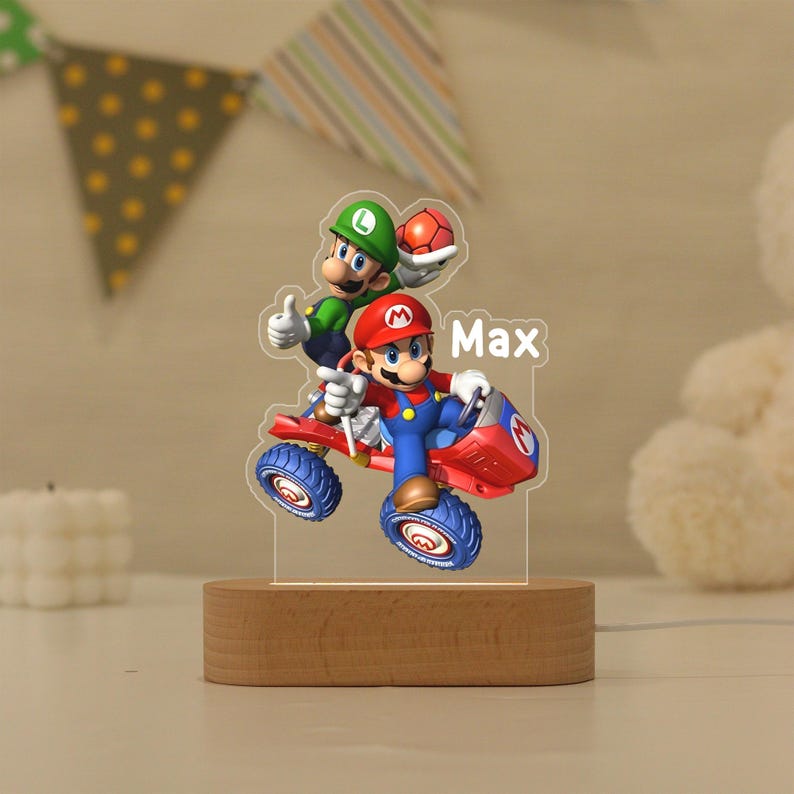 May include: A custom acrylic night light featuring a colourful graphic of Mario driving a red kart with Luigi standing behind him. The name Max is printed in white text on the right side. The acrylic piece is mounted on a smooth oval wooden base.