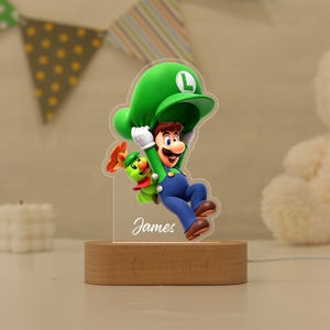 May include: An acrylic night light featuring a colourful graphic of Luigi holding a green caterpillar-like creature, set on a light wood base. The design includes the name James in white script text. The character wears a green hat with an L.