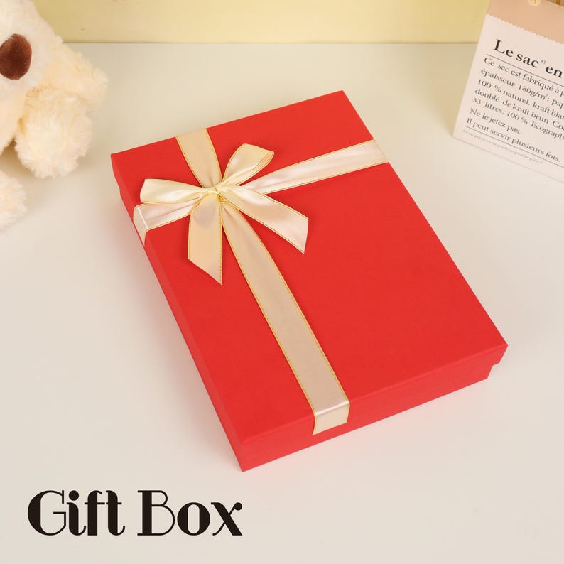 May include: A rectangular red gift box featuring a smooth matte finish and a decorative cream-coloured satin ribbon tied in a bow across the top. The ribbon is positioned off-centre, creating a clean and structured presentation for the box.
