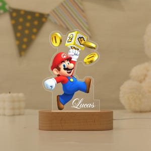 May include: A custom acrylic night light featuring a colourful graphic of Mario jumping towards a yellow question block and gold coins. The clear acrylic panel is personalised with the name Lucas in white script and sits on a light wood base.