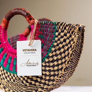 May include: A hand-woven basket with a brown leather handle and a tag reading "Vetavera Collectibles Authentically Handcrafted." The basket has a colourful design with pink, green, and black woven patterns. The handle is wrapped with brown leather.