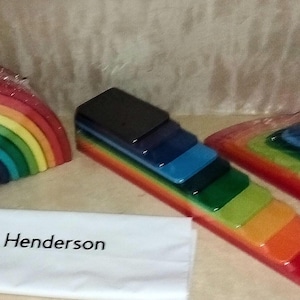 May include: A selection of wooden rainbow stacking toys in a range of sizes and colours, including red, orange, yellow, green, blue, and purple. The toys are packaged in clear plastic. A white paper with the name "Henderson" is in front.