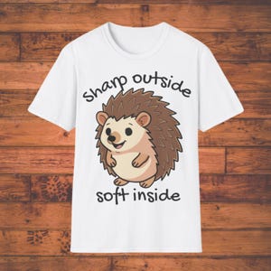 Puede incluir: Una taza de cerámica blanca con un asa negra y el texto "But First, Coffee" en negrita negra. La taza se encuentra sobre un fondo borroso, sugiriendo un ambiente acogedor.