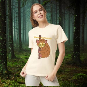 May include: Cream-colored t-shirt featuring a cartoon tarsier clinging to a branch. The tarsier is brown with large yellow eyes. The text "Always Night Mode" is above the tarsier. The background is a forest scene.