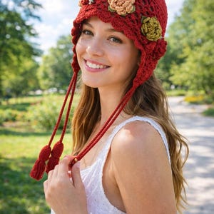 Könnte beinhalten: Ein roter gehäkelter Hut mit floralen Verzierungen und Quasten. Der Hut hat einen Glockenstil mit einem gewellten Rand und ist mit gehäkelten Blumen in Beige und Grün verziert. Quasten hängen auf beiden Seiten des Gesichts herunter.