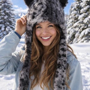 May include: A faux fur hat with ear flaps and long ties, featuring a black and gray spotted pattern. The hat has black ear tips and is worn in a snowy outdoor setting. The person is wearing a light blue jacket.