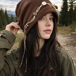May include: A brown beanie hat with decorative detailing. The hat has a cream-coloured band with wooden beads and a large, white shell-like embellishment. The person in the image is wearing an olive green jacket.