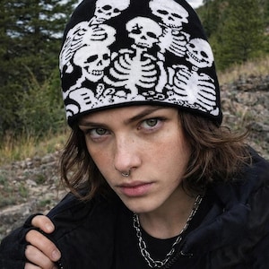 May include: Black beanie featuring a white skeleton design. The hat is worn by a person with a silver chain necklace and a black jacket. The background is a blurred outdoor scene with trees and rocks.