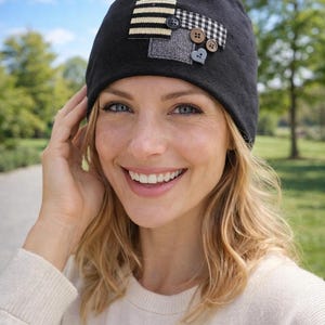 Könnte beinhalten: Schwarze Mütze mit dekorativen Aufnähern und Knöpfen. Die Aufnäher umfassen ein Streifendesign, ein Karomuster und ein graues Quadrat. Die Knöpfe sind in verschiedenen Farben und Größen. Die Mütze wird von einer Person mit blondem Haar getragen.