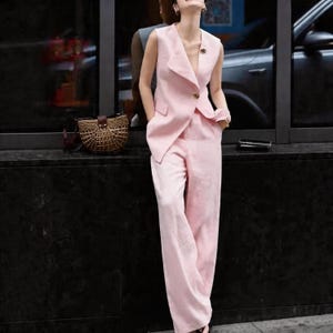 May include: A light pink sleeveless suit, featuring a tailored vest with a single gold button and matching wide-leg pants. A small, decorative pin adorns the vest. A woven handbag with a leather strap is visible in the background.