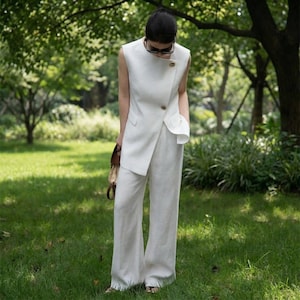 May include: A white linen sleeveless top with a wrap-style front, paired with matching wide-leg pants. The top features a gold button closure and a small pocket. The outfit is worn outdoors in a grassy area, with a brown handbag.