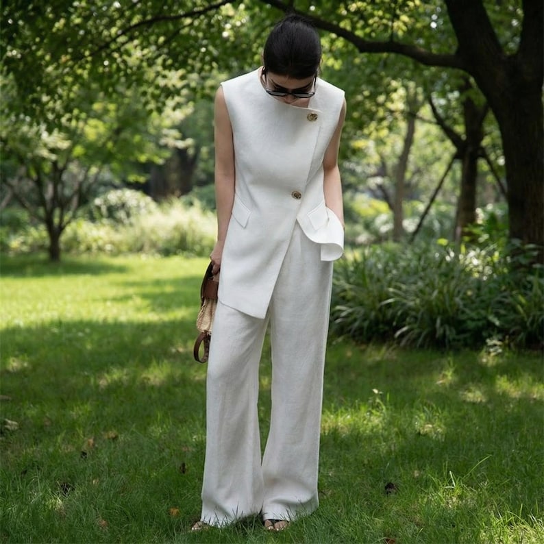 May include: A white linen sleeveless top with a wrap-style front, paired with matching wide-leg pants. The top features a gold button closure and a small pocket. The outfit is worn outdoors in a grassy area, with a brown handbag.