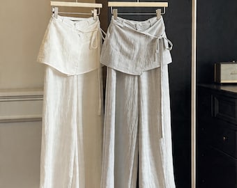 May include: Two sets of linen pants and wrap tops hang on a clothing rack. One set is white, the other is light gray. The tops have an asymmetrical wrap design with a button closure and a matching tie. The pants are wide-legged.