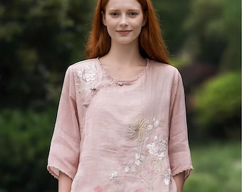 May include: A light pink linen top with three-quarter sleeves and floral embroidery. The top features a round neckline with delicate trim and a button closure. The embroidery includes white and beige flowers and leaves, adding a touch of elegance to the garment.