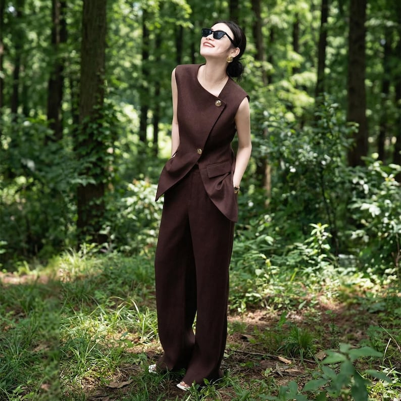 May include: A brown sleeveless vest with a single button closure and matching wide-leg pants. The outfit is a solid color and appears to be made of a soft fabric. The vest has a unique asymmetrical design. The person is wearing sunglasses.