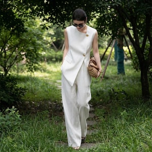 May include: A white linen suit, consisting of a sleeveless top with a diagonal closure and wide-leg pants. The top features a gold button detail. A woven handbag is carried. The outfit is worn outdoors in a natural setting.