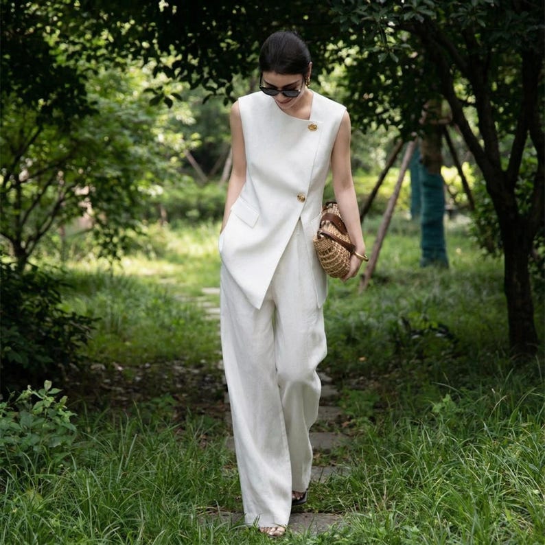 May include: A white linen suit, consisting of a sleeveless top with a diagonal closure and wide-leg pants. The top features a gold button detail. A woven handbag is carried. The outfit is worn outdoors in a natural setting.