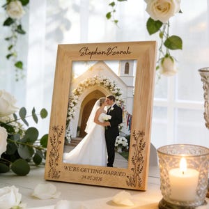 Peut inclure: Un cadre photo en bois clair contenant une photo de mariage. Le cadre est gravé des noms "Stephan & Sarah" et de la date "29.05.2025". La photo montre un couple s'embrassant devant une église.