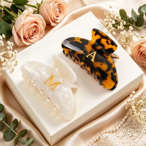 Op de afbeelding: Twee haarklemmen met een glanzende afwerking. De ene klem is wit met een parelmoerachtige textuur en een gouden letter M, terwijl de andere bruin en zwart schildpadpatroon heeft met een gouden letter A. Beide klemmen hebben een gebogen ontwerp.