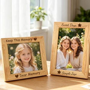 Puede incluir: Dos marcos de fotos de madera clara con texto grabado. El marco horizontal tiene el texto Keep This Memory y Dear Memory con corazones. El marco vertical tiene el texto Sweet Days y Simple Joys con estrellas.