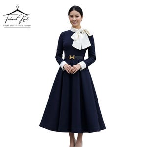 Puede incluir: Un vestido azul marino con un lazo blanco y puños blancos. El vestido tiene un cinturón negro con hebilla dorada. El vestido es de manga larga y tiene una falda amplia. El logo de Tailored Knot está en la esquina superior izquierda.