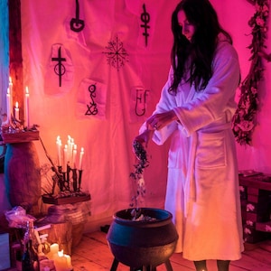 May include: A person in a white robe pours herbs into a black cauldron, surrounded by candles and mystical symbols. The scene is illuminated with pink and purple lighting, creating an atmospheric setting. The cauldron sits on a black stand.