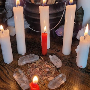 May include: A selection of lit white and red candles, crystals, and herbs arranged on a wooden surface. A black cauldron and other spiritual items are in the background. The candles are of varying heights, with flames casting a warm glow.