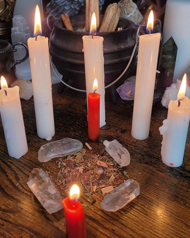 May include: A selection of lit white and red candles, crystals, and herbs arranged on a wooden surface. A black cauldron and other spiritual items are in the background. The candles are of varying heights, with flames casting a warm glow.