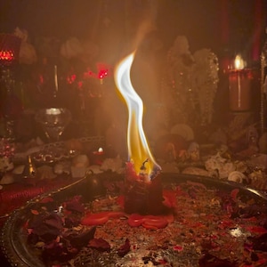 May include: A burning red candle with a tall, bright flame. The candle is in a silver tray with dried rose petals and ash. The background is dimly lit with other candles and decorative items, creating a mystical atmosphere.