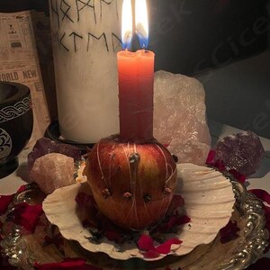 May include: A burning red candle is placed on a decorated apple, set on a seashell within a silver tray. A white candle with runes is behind the red candle. Rose petals and crystals surround the arrangement.