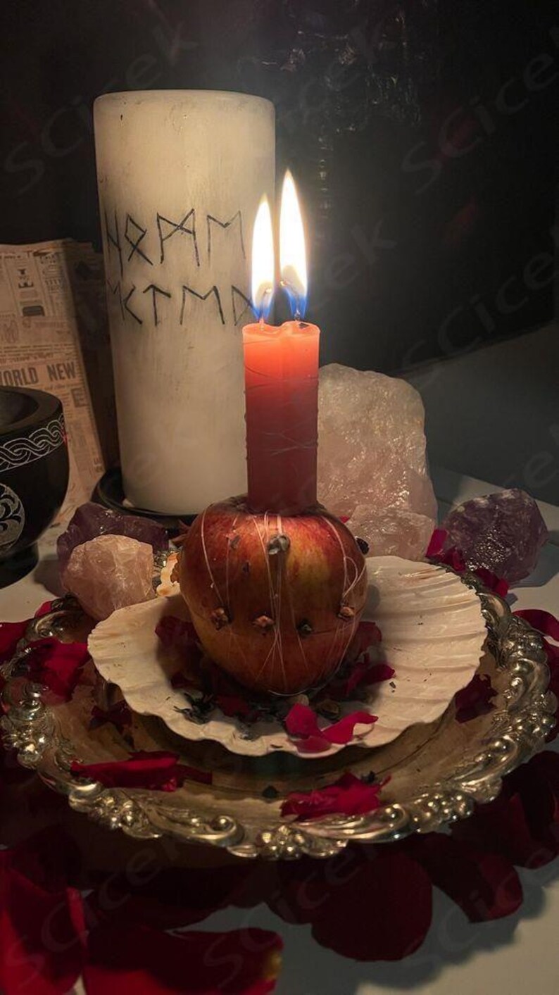 May include: A burning red candle is placed on a decorated apple, set on a seashell within a silver tray. A white candle with runes is behind the red candle. Rose petals and crystals surround the arrangement.