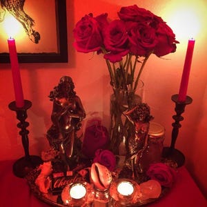 May include: A decorative arrangement with red roses in a glass vase, two red candles in black holders, and bronze statues. A mirrored tray holds lit candles, a seashell, and a small box of chocolate. The scene is illuminated by a warm, red light.