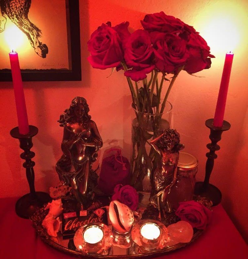 May include: A decorative arrangement with red roses in a glass vase, two red candles in black holders, and bronze statues. A mirrored tray holds lit candles, a seashell, and a small box of chocolate. The scene is illuminated by a warm, red light.