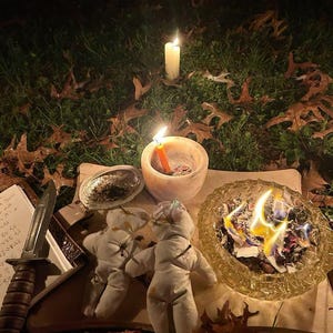 May include: A nighttime ritual scene with two lit candles, a small bowl with a burning candle, two cloth dolls, a knife, and a glass bowl with burning paper. These items are arranged on a wooden surface, surrounded by fallen leaves.
