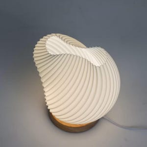 Könnte beinhalten: Eine weiße Tischleuchte mit einem geschwungenen, muschelartigen Schirm mit wiederkehrender Rippenstruktur. Der Schirm ist auf einem runden Holzsockel montiert und strahlt ein weiches, diffuses Licht aus, das eine skulpturale Form bildet.